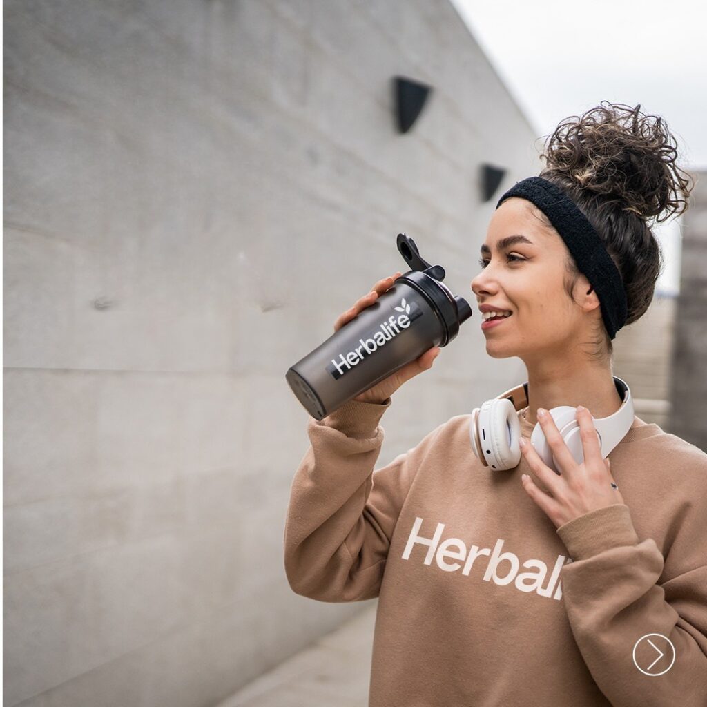eune femme souriante en sweat beige Herbalife, tenant un shaker noir Herbalife et portant un casque audio autour du cou