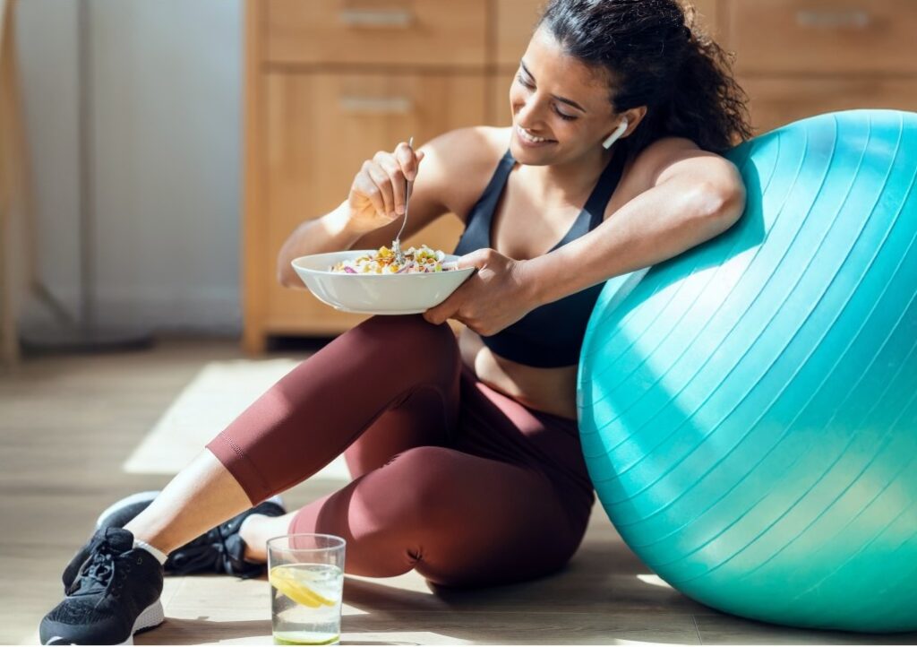 Femme en tenue de sport assise au sol, savourant un repas équilibré à côté d’un ballon de fitness et d’un verre d’eau citronnée